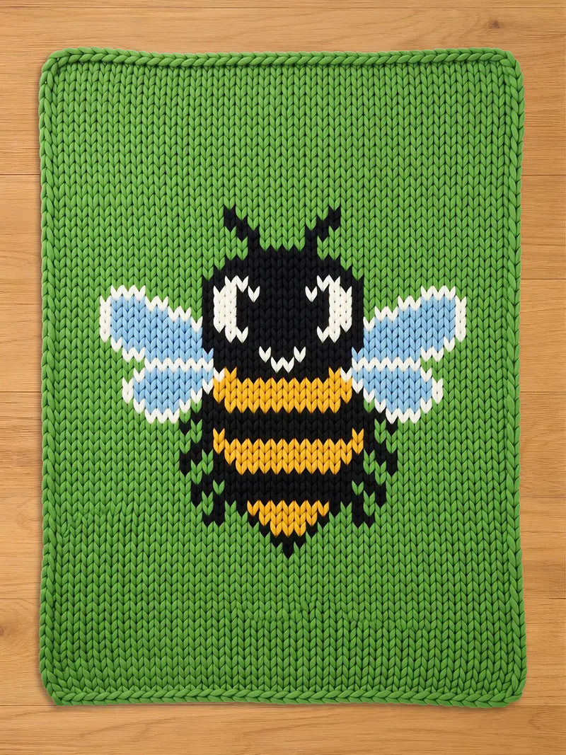 A knitted green blanket with a large, central design of a cartoon bee. The bee features a black head and body stripes, yellow stripes, and light blue wings. The blanket has a green border.