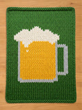 A chunky knit blanket in green featuring a large, textured pattern of a beer mug filled with yellow beer and white foam, displayed flat on a wooden surface