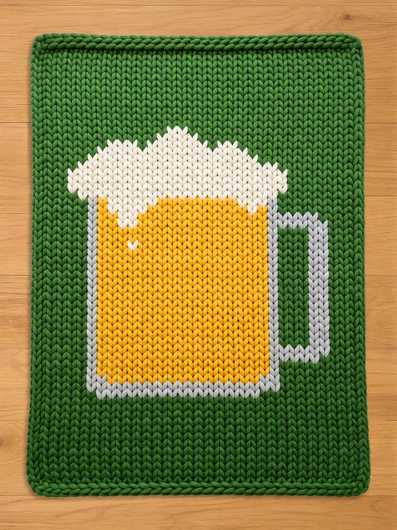 A chunky knit blanket in green featuring a large, textured pattern of a beer mug filled with yellow beer and white foam, displayed flat on a wooden surface