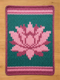 A knitted chunky blanket with a lotus flower design. The blanket features a teal background with a pink and purple lotus flower in the center. The border and horizontal stripes are pink and purple.