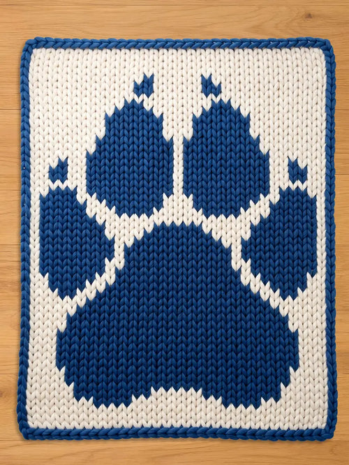 a chunky knitted blanket featuring a large blue paw print on a white background. The paw print is centered, with a blue border around the edge. The blanket is made of thick yarn.