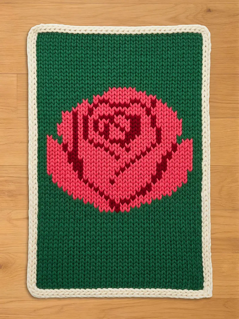 A knitted rectangular chunky blanket featuring a large pink rose on a green background. The rose is detailed with darker pink shading. The blanket has a cream-colored border. The blanket is made of a thick, textured yarn.