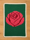 A knitted rectangular chunky blanket featuring a large pink rose on a green background. The rose is detailed with darker pink shading. The blanket has a cream-colored border. The blanket is made of a thick, textured yarn.