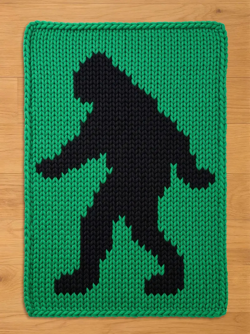 A knitted green and black blanket featuring a silhouette of a Bigfoot. The Bigfoot figure is black and stands out against the green background. The blanket is rectangular with a thick, knitted border.