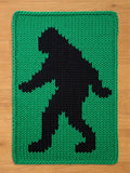 A knitted green and black blanket featuring a silhouette of a Bigfoot. The Bigfoot figure is black and stands out against the green background. The blanket is rectangular with a thick, knitted border.