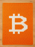 An orange knitted blanket featuring a large white Bitcoin symbol. The blanket has a textured appearance due to the knitted design. The Bitcoin symbol is centered on the blanket.