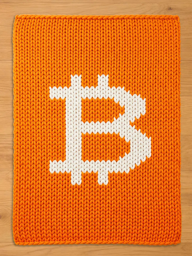 An orange knitted blanket featuring a large white Bitcoin symbol. The blanket has a textured appearance due to the knitted design. The Bitcoin symbol is centered on the blanket.
