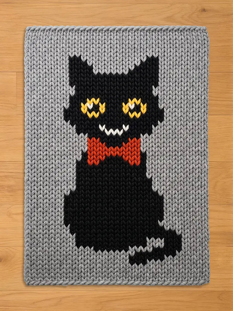 Chunky knit blanket in light gray featuring a knitted black cat pattern with a red bow tie and yellow eyes.