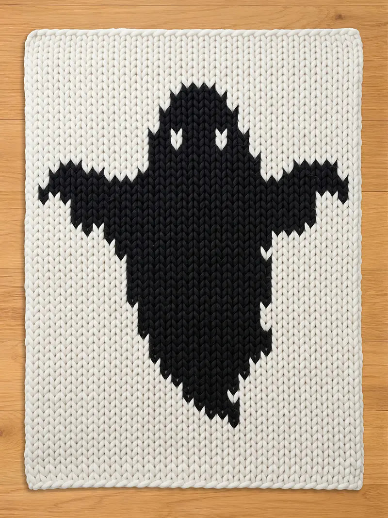 A rectangular chunky knit blanket featuring a large, centered black ghost silhouette against a cream-colored background. The textile displays a prominent, thick yarn texture with a consistent stitch pattern throughout the entire piece.