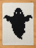 A rectangular chunky knit blanket featuring a large, centered black ghost silhouette against a cream-colored background. The textile displays a prominent, thick yarn texture with a consistent stitch pattern throughout the entire piece.
