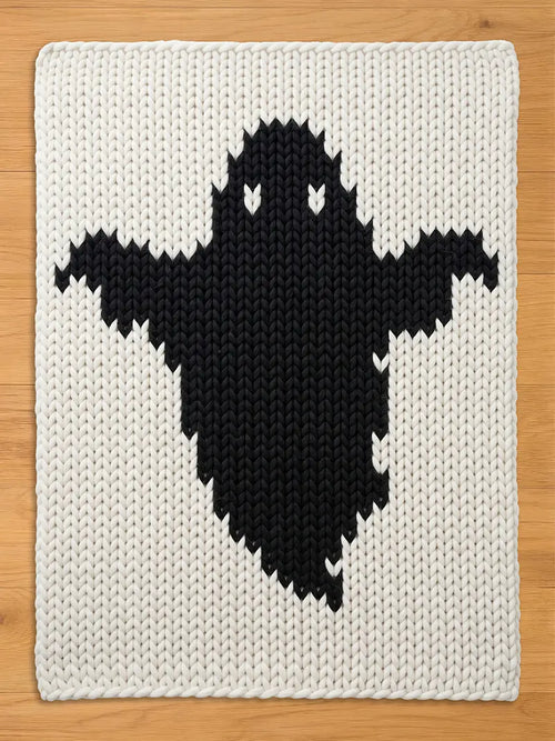 A rectangular chunky knit blanket featuring a large, centered black ghost silhouette against a cream-colored background. The textile displays a prominent, thick yarn texture with a consistent stitch pattern throughout the entire piece.