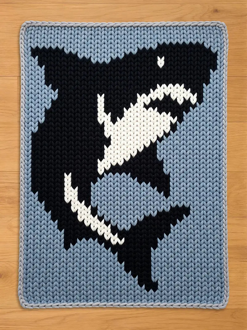 Chunky knit blanket featuring a black, white, and grey shark design on a light blue background, laid flat on a wooden floor.