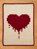 A knitted rectangular chunk knit blanket with a cream-colored background and a large, pixelated red heart design. The heart has a dripping effect at the bottom. The blanket is trimmed with a red border.