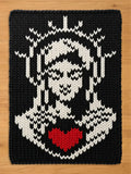 A chunky knit blanket featuring a black and white knit pattern of the Blessed Mother with a vibrant red heart in the center, displayed flat on a wooden surface.