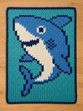 A chunky knit blanket featuring a woven design of a friendly blue and white shark. The blanket uses thick, oversized yarn in shades of teal, royal blue, and cream, knit in a distinct stockinette stitch pattern. It is laid flat on a light wooden floor background.