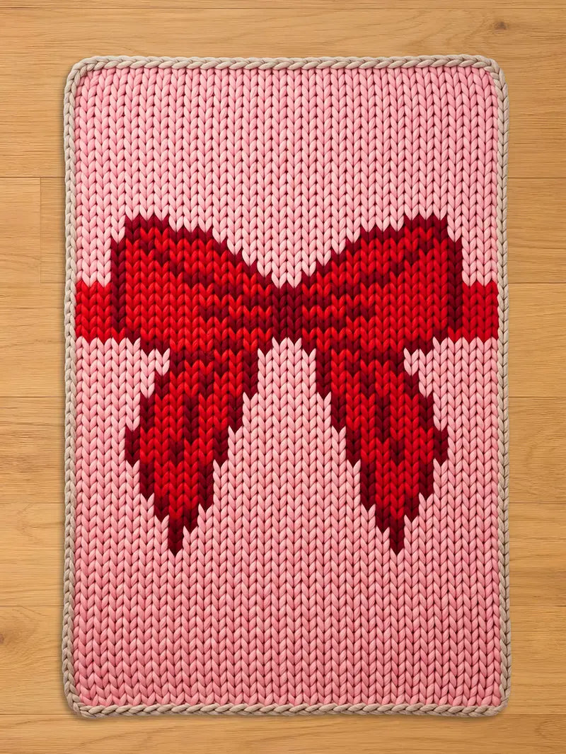  A knitted chunky blanket with a large red bow design on a pink background. The bow is a darker shade of red, and the rug has a light beige border. The blanket is rectangular and appears to be handmade.