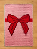  A knitted chunky blanket with a large red bow design on a pink background. The bow is a darker shade of red, and the rug has a light beige border. The blanket is rectangular and appears to be handmade.