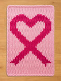 Light pink chunky knit blanket featuring a large, dark pink breast cancer awareness ribbon shaped like a heart in the center, displayed on a wooden floor background.