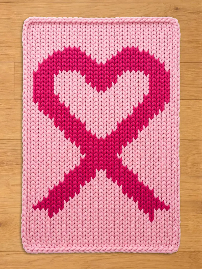 Light pink chunky knit blanket featuring a large, dark pink breast cancer awareness ribbon shaped like a heart in the center, displayed on a wooden floor background.