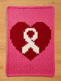 A knitted pink blanket featuring a red heart with a white ribbon in the center. The blanket is rectangular with a thick, textured knit pattern. The heart and ribbon design is a symbol of awareness.