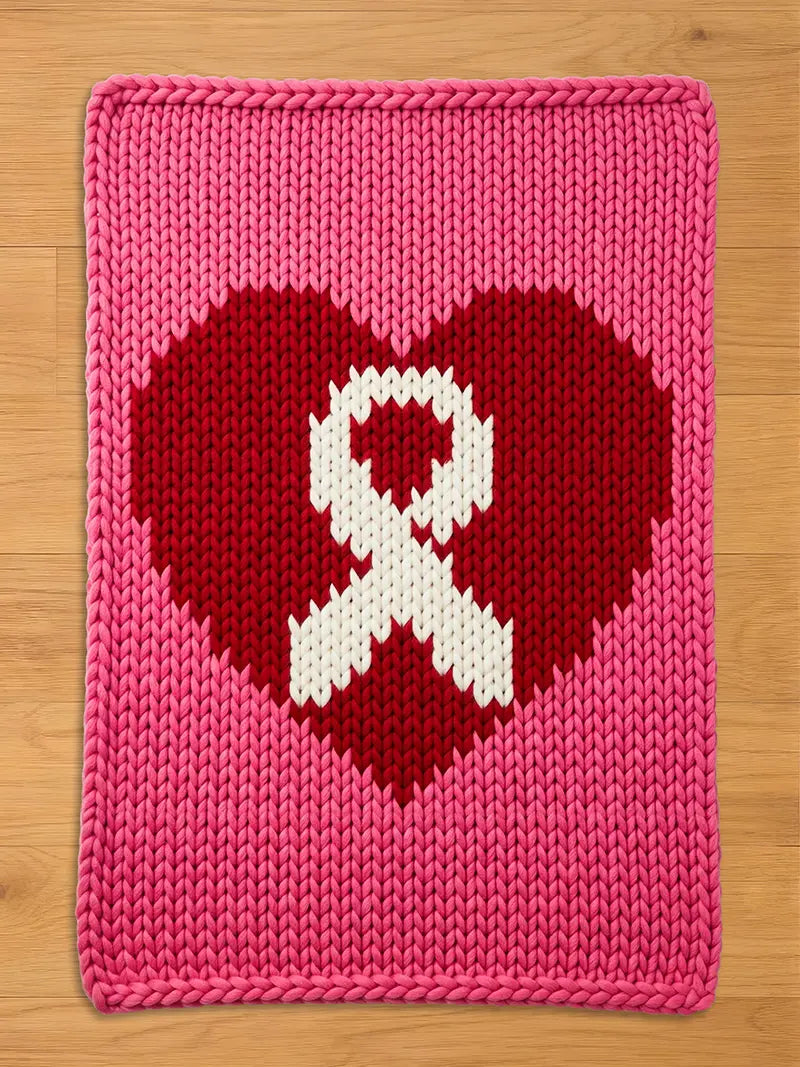 A knitted pink blanket featuring a red heart with a white ribbon in the center. The blanket is rectangular with a thick, textured knit pattern. The heart and ribbon design is a symbol of awareness.