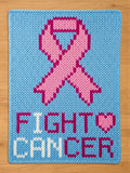 A knitted chunky blanket in light blue features a pink ribbon and the words "FIGHT CANCER" in white and pink. The heart symbol is pink. The design is a statement of support and awareness.