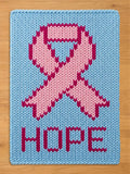 A rectangular light blue knitted blanket featuring a central pink awareness ribbon design with a darker pink outline. The word HOPE is knitted in dark pink capital letters at the bottom of the blanket, centered below the ribbon.