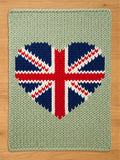 A rectangular sage green chunky knit blanket featuring a large, centered heart-shaped design. The heart is patterned with the Union Jack flag, rendered in deep navy blue, bright red, and white yarn, creating a bold, textured contrast.