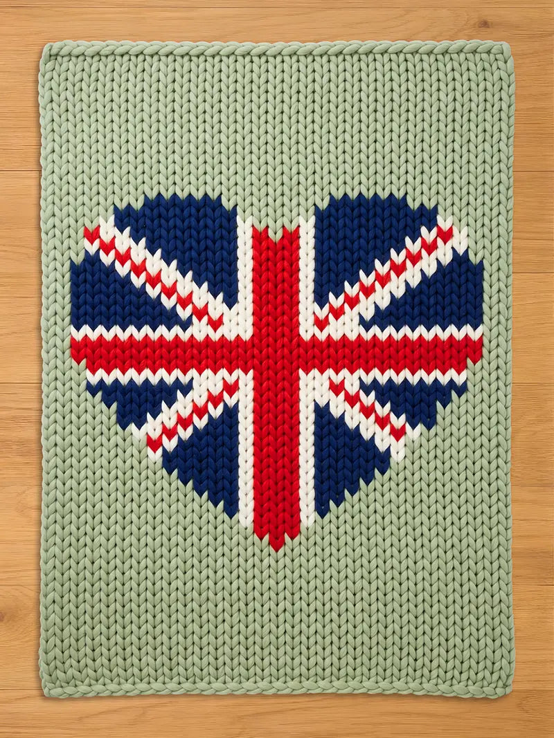 A rectangular sage green chunky knit blanket featuring a large, centered heart-shaped design. The heart is patterned with the Union Jack flag, rendered in deep navy blue, bright red, and white yarn, creating a bold, textured contrast.