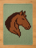 A knitted rectangular chunky blanket featuring a horse head design. The horse head is rendered in shades of brown against a light green background. The blanket is made of a thick, textured yarn.