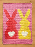 A pink knitted blanket featuring two bunny figures. One bunny is red, the other is yellow, both with white heart-shaped details. The blanket is made of thick yarn.