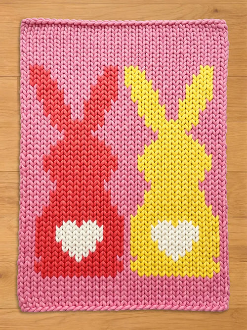A pink knitted blanket featuring two bunny figures. One bunny is red, the other is yellow, both with white heart-shaped details. The blanket is made of thick yarn.