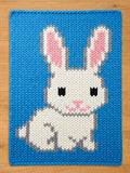 A knitted blue square chunky blanket featuring a white bunny design. The bunny has pink inner ears, gray accents, and black eyes. The blanket is made of a thick, textured yarn.