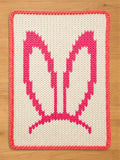 Chunky knit blanket featuring a pink bunny ears pattern on a cream white background with a pink braided border.