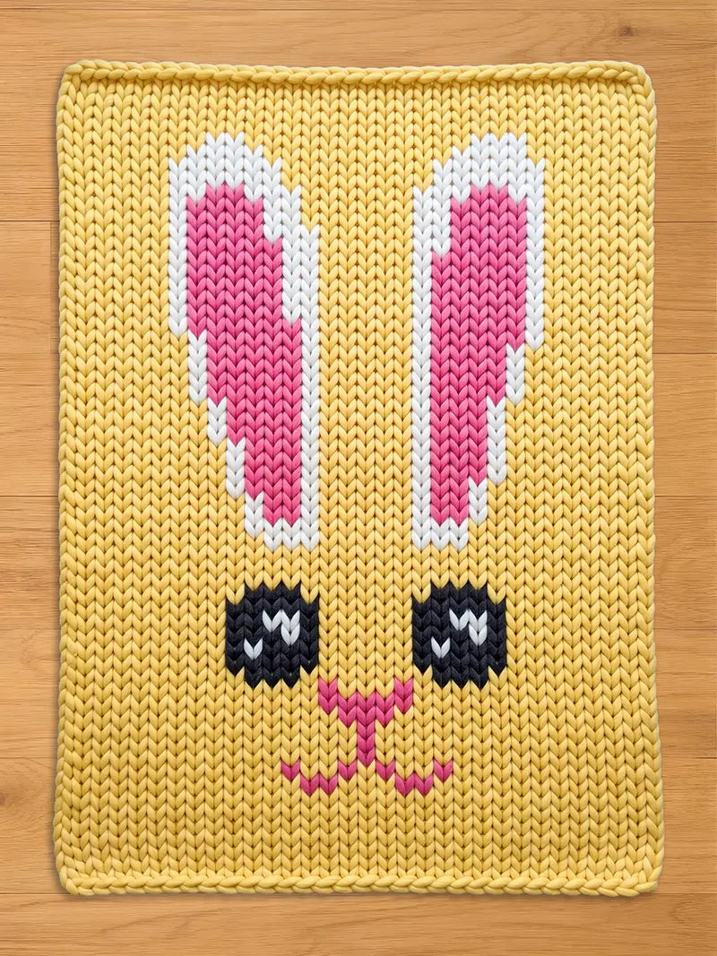 Yellow chunky knit blanket featuring a large knitted bunny face pattern with pink and white ears, large black eyes, and a pink nose and mouth, displayed flat on a wooden surface.