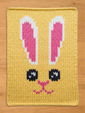 Yellow chunky knit blanket featuring a large knitted bunny face pattern with pink and white ears, large black eyes, and a pink nose and mouth, displayed flat on a wooden surface.