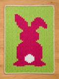 A chunky knit blanket featuring a vibrant pink bunny silhouette with a white fluffy tail on a lime green background, finished with a cream-colored braided border.