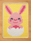 A knitted Easter-themed chunky knit blanket featuring a pink bunny emerging from a white egg against a yellow background. The bunny has pink ears and a smiling face. The border is cream-colored.