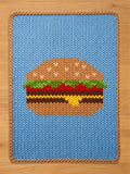 A knitted blue rectangular chunky blanket with a brown border. The blanket features a large, detailed burger design in shades of brown, red, green, and yellow. The burger is centered on the blanket, creating a fun, food-themed accent.