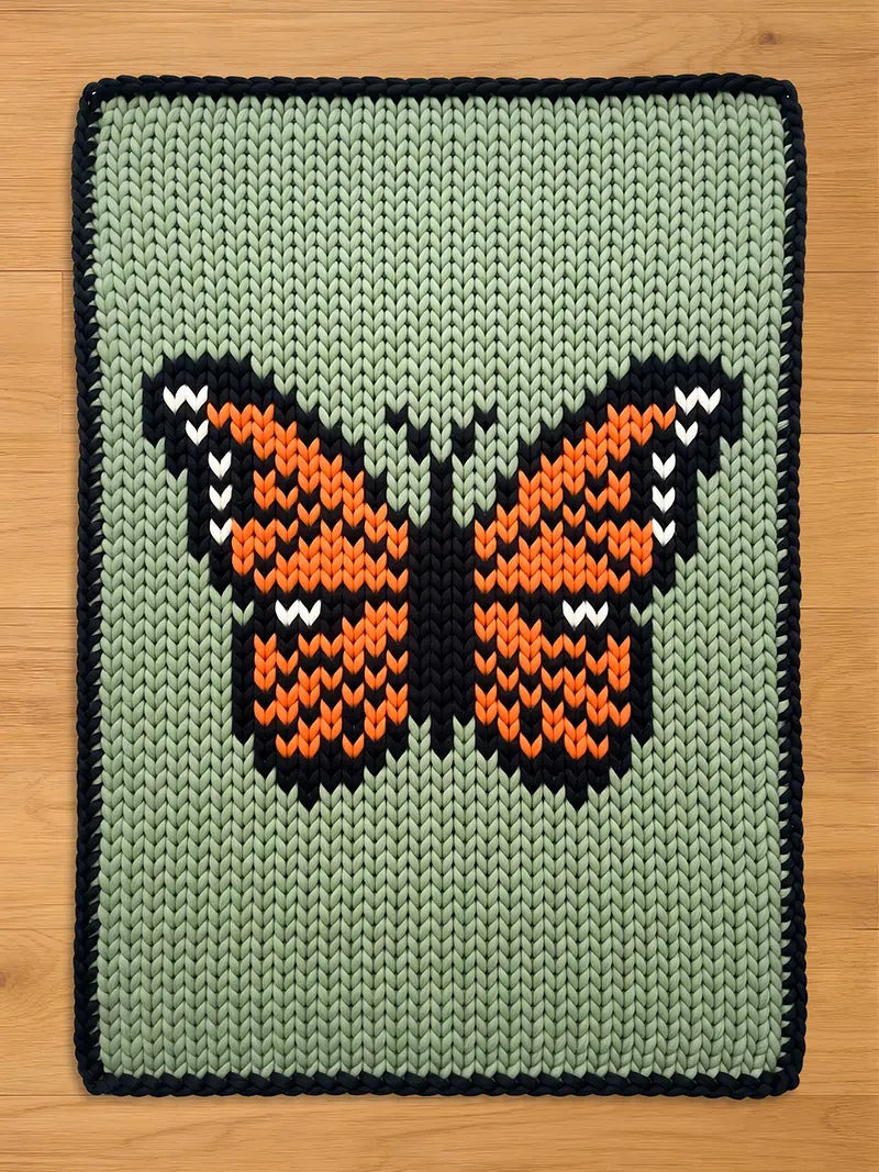 A knitted chunky blanket featuring a butterfly design. The butterfly has orange and black wings with white accents, set against a light green background. The blanket has a dark blue border.