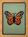 A knitted chunky blanket featuring a butterfly design. The butterfly has orange and black wings with white accents, set against a light green background. The blanket has a dark blue border.