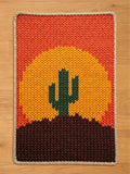 A knitted chunky blanket featuring a desert sunset scene. The design includes a green cactus on a brown hill, a yellow and orange sun, and an orange and red sky. The edges are finished with a beige border.