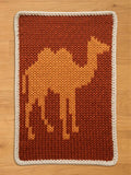 A knitted camel design on a rectangular chunky blanket. The camel is in shades of orange and yellow against a brown background. The blanket has a white border. This handmade item is suitable for a nursery or children's room.