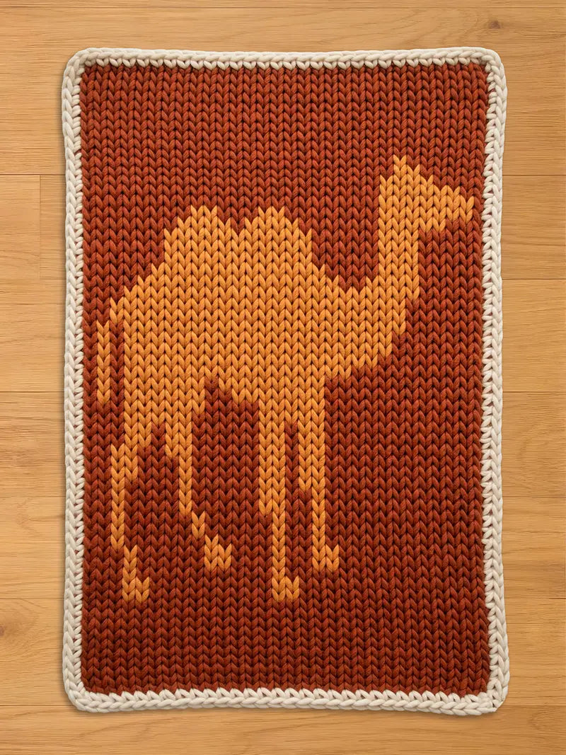 A knitted camel design on a rectangular chunky blanket. The camel is in shades of orange and yellow against a brown background. The blanket has a white border. This handmade item is suitable for a nursery or children's room.