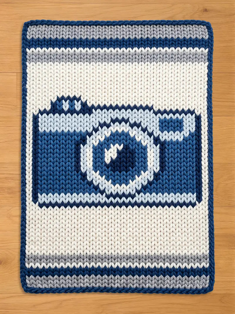Chunky knit blanket featuring a woven dark blue, light blue, and grey vintage camera design on a cream-white background with a dark blue border, laid flat on a wooden floor.
