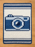 Chunky knit blanket featuring a woven dark blue, light blue, and grey vintage camera design on a cream-white background with a dark blue border, laid flat on a wooden floor.