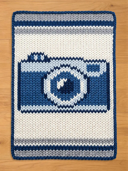 Chunky knit blanket featuring a woven dark blue, light blue, and grey vintage camera design on a cream-white background with a dark blue border, laid flat on a wooden floor.