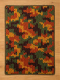 A chunky knit blanket featuring a camouflage pattern in autumn colors, including forest green, deep brown, burnt orange, and mustard yellow, laid flat on a light wood floor.