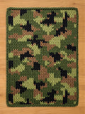 A chunky knit blanket featuring a classic camouflage pattern in shades of olive green, light green, tan, and black. The image shows the blanket flat on a light wood floor, highlighting its thick, braided border and texture.