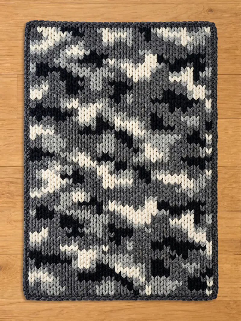 A chunky knit blanket in a winter camouflage pattern with black, grey, and cream yarn, displayed flat on a light wood floor.